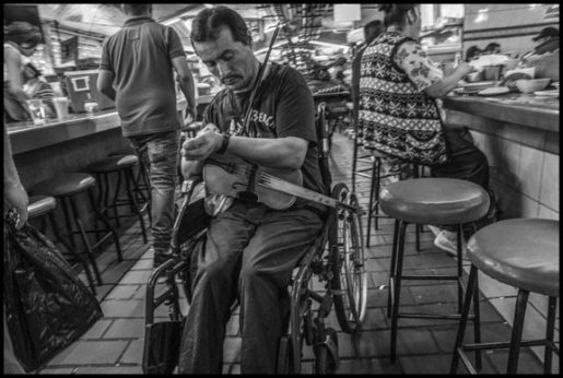 A violinist in his wheelchair checks his watch after getting a bite to eat.  Maybe he has a spot in the market to play for only a certain time, and can't be late. | David Bacon